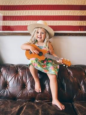 Little girl sat on the back of a leather sofa wearing a hat, holding a small guitar